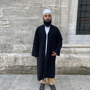 May include: A black open-front robe worn over a white shirt and tan trousers, paired with a white skullcap and dark gray sneakers. The robe features long sleeves and a simple, unadorned design, providing a layered look against a stone wall.