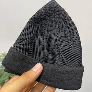 May include: A black kufi cap featuring a textured knit pattern with vertical diamond shapes and a solid, smooth fabric band at the base. The band is decorated with subtle, horizontal wavy stitching that runs along the entire circumference.