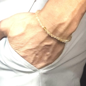 May include: A delicate bracelet made of small, clear beads with a subtle golden hue. The bracelet is worn on a person's wrist.