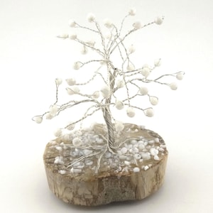 Puede incluir: Una escultura de árbol decorativa hecha a mano. El árbol presenta un tronco y ramas de alambre plateado adornados con cuentas esféricas blancas. Se asienta sobre una base de madera natural de color marrón claro, creando un artículo de decoración único.
