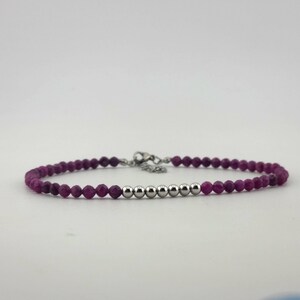 May include: A delicate bracelet featuring small, faceted, deep magenta beads interspersed with a series of polished silver-colored beads. The bracelet has a silver-toned clasp and chain for adjustable sizing. Jewelry.