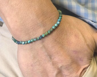 May include: A beaded bracelet with small, round, turquoise-colored beads. The bracelet is worn on a wrist, with the beads displaying shades of blue and green. The background includes a light blue and white checkered shirt.