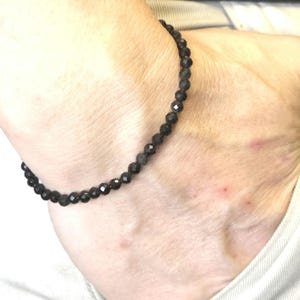 May include: A black beaded bracelet with faceted beads. The bracelet is worn on the wrist and is made of small, dark beads strung together. The beads have a shiny, reflective surface.