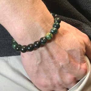Puede incluir: Una pulsera de cuentas compuesta por piedras esféricas de color verde oscuro liso, con una cuenta única jaspeada en amarillo y verde. Las cuentas están ensartadas en un patrón circular, creando un bucle continuo de esferas de piedra pulida.