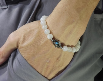 May include: White beaded bracelet with a silver hamsa hand charm featuring a blue eye. The bracelet is on a person's wrist.