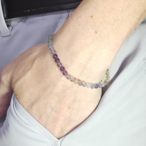 May include: A bracelet made of small, round, multi-colored beads. The beads are shades of purple, blue, and green. The bracelet is worn on a person's wrist.