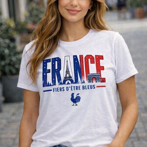 May include: White t-shirt with the word "FRANCE" in blue and red, featuring the Eiffel Tower and Arc de Triomphe. Below the text is "FIERS D'&Ecirc;TRE BLEUS" and a blue rooster graphic. The shirt is worn with blue jeans.
