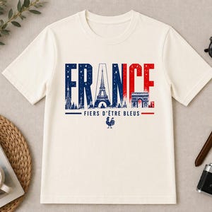 May include: Cream-colored t-shirt with the word "FRANCE" in blue and red, featuring the Eiffel Tower and Arc de Triomphe. The text "FIERS D'&Ecirc;TRE BLEUS" and a rooster graphic are below. Accessories include sunglasses, a watch, and a camera.