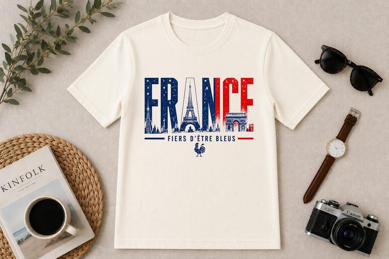 May include: Cream-colored t-shirt with the word "FRANCE" in blue and red, featuring the Eiffel Tower and Arc de Triomphe. The text "FIERS D'&Ecirc;TRE BLEUS" and a rooster graphic are below. Accessories include sunglasses, a watch, and a camera.