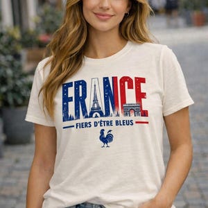 May include: Off-white t-shirt with the word "FRANCE" in blue and red, featuring the Eiffel Tower and Arc de Triomphe. Below, the text "FIERS D'&Ecirc;TRE BLEUS" and a blue rooster graphic. The shirt is worn by a person.