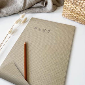 Könnte beinhalten: Ein beigefarbenes kariertes Notizbuch mit handgeschriebenen Matheaufgaben und einem braunen Bleistift. Das Notizbuch ist geöffnet und zeigt eine zweite Seite darunter. Trockenblumen und ein Weidenkorb befinden sich im Hintergrund.