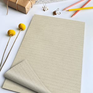 Könnte beinhalten: Ein beigefarbenes, liniertes Notizbuch mit leicht strukturierter Oberfläche. Das Notizbuch ist teilweise geöffnet und zeigt die Innenseiten. Zwei gelbe Bleistifte, zwei goldene Büroklammern und zwei gelbe Trockenblumen sind um das Notizbuch angeordnet.