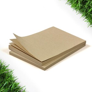 Könnte beinhalten: Ein Stapel quadratischer, hellbrauner Papierblöcke mit ein paar angehobenen Seiten, vor weißem Hintergrund. Das Papier hat ein strukturiertes Aussehen. Grünes Gras befindet sich in der oberen rechten und unteren linken Ecke.