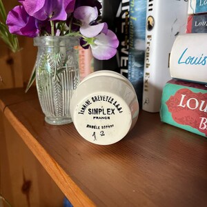 May include: An off-white ceramic jar with black text reading "SIMPLEX FRANCE" sits on a wooden shelf. A small glass vase with purple and white flowers is next to the jar. Books are visible in the background.
