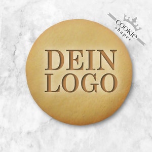 以下が含まれることがあります： 「DEIN LOGO」の文字が濃い茶色のフォントでエンボス加工された、丸い薄茶色のクッキー。クッキーの表面はわずかに不均一で、白い大理石の背景に置かれています。「cookie shaper」の文字が灰色のリボンに印刷されています。