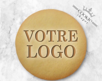 May include: A round, light brown cookie with the words "VOTRE LOGO" imprinted on its surface. The cookie has a smooth texture and is set against a marble background. The text "cookie shaper" is also visible.