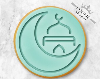 May include: A light blue cookie cutter shaped like a crescent moon with a mosque inside. The cookie cutter is on a white marble background.