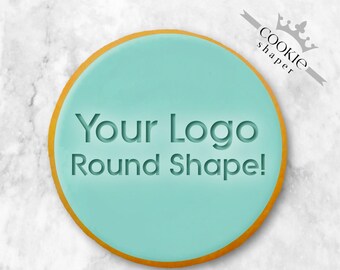 May include: A round cookie with a light teal frosting and a gold edge. The words "Your Logo Round Shape!" are embossed into the frosting. The cookie is on a white marble surface. The cookie is a product of Cookie Shaper.