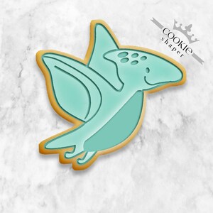 Peut inclure: Un emporte-pièce en forme de dinosaure volant de couleur bleu turquoise avec une grande envergure et une petite tête avec des yeux. L'emporte-pièce est en métal et a une finition dorée.