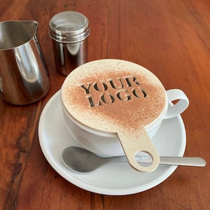 K&ouml;nnte beinhalten: Eine wei&szlig;e Kaffeetasse mit Untertasse, L&ouml;ffel und Schablone mit den Worten "YOUR LOGO", best&auml;ubt mit Kakaopulver. Ein Edelstahl-Milchk&auml;nnchen und ein Shaker befinden sich ebenfalls auf der Holzoberfl&auml;che.