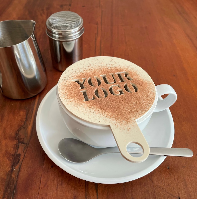 K&ouml;nnte beinhalten: Eine wei&szlig;e Kaffeetasse mit Untertasse, L&ouml;ffel und Schablone mit den Worten "YOUR LOGO", best&auml;ubt mit Kakaopulver. Ein Edelstahl-Milchk&auml;nnchen und ein Shaker befinden sich ebenfalls auf der Holzoberfl&auml;che.