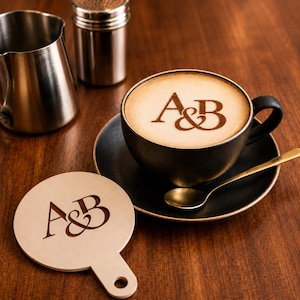 Può includere: Una tazza da caffè in ceramica nera con latte art che raffigura le lettere A&B, appoggiata su un piattino nero abbinato con un cucchiaino dorato. Accanto si trova uno stencil circolare in legno chiaro con le lettere A&B ritagliate.