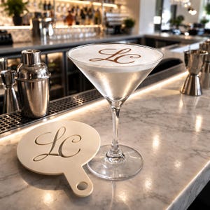 Può includere: Un cocktail martini in un bicchiere trasparente con una schiuma bianca decorata da un monogramma LC in corsivo marrone, accompagnato da uno stencil bianco rotondo con manico e un motivo LC ritagliato, appoggiato su una superficie di marmo.