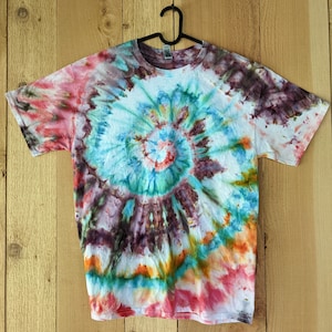 May include: A tie-dye t-shirt with a swirling pattern in shades of red, orange, yellow, green, blue, and purple. The shirt is hanging on a black hanger against a wooden background.