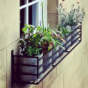 May include: Black metal window box planter with five potted plants. The plants include green foliage, lavender, and pink flowers. The planter is mounted on a white brick wall.