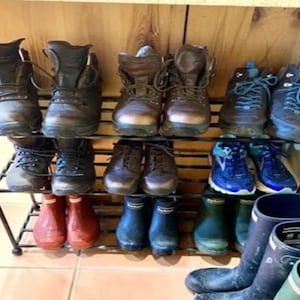 May include: A black metal shoe rack displaying an assortment of footwear. Boots, walking boots, and wellington boots in brown, black, red, and blue are organised on the shelves. The rack is placed against a wooden wall.