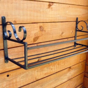May include: A black metal shelf with decorative scrollwork brackets mounted on a wooden wall. The shelf has a simple, rectangular design and is ideal for displaying small items.