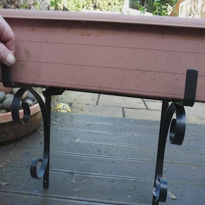 May include: A brown rectangular planter box supported by black wrought iron scrollwork brackets. The brackets are fixed to a wooden deck. A hand is holding the planter box.