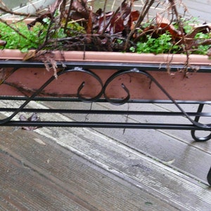 May include: A black metal plant stand with a decorative scrolled design. The stand has a rectangular planter box with a light brown finish. The planter box is filled with green plants and brown twigs.