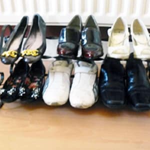 May include: A black metal shoe rack with two tiers holds a variety of shoes, including black leather shoes, white trainers, and black and white patterned shoes.