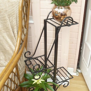 May include: Black metal plant stand with three shelves. The stand features a decorative scroll design. A potted plant is on the top shelf.
