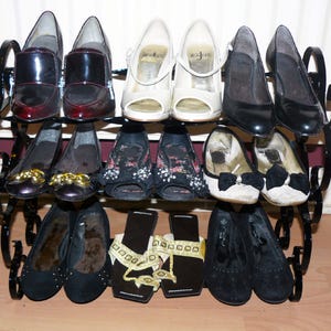 May include: A black metal shoe rack with three tiers holds a variety of shoes, including black, brown, and white heels, black flats, and black sandals.