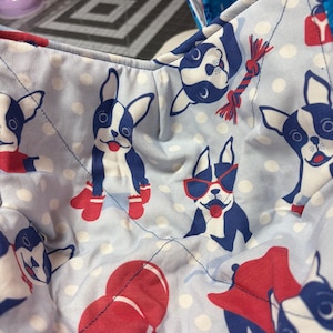May include: Light blue fabric bowl with a pattern of Boston Terrier dogs in various poses. The dogs are in blue and white with red accents, including boots, sunglasses, and balloons. White polka dots are scattered across the fabric.