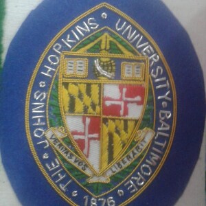 May include: A blue and gold embroidered patch with the text "The Johns Hopkins University Baltimore 1876" and a coat of arms featuring a shield with a red cross on a white background, a yellow and black striped background, and a green and gold background.