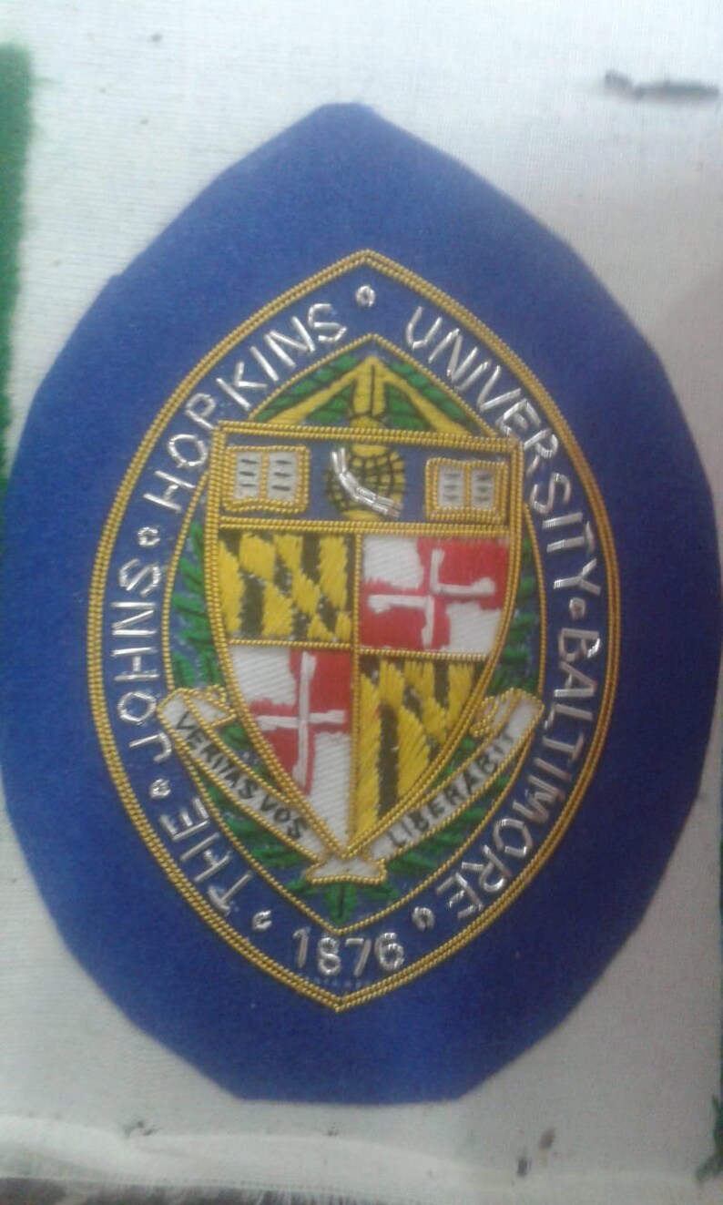 May include: A blue and gold embroidered patch with the text "The Johns Hopkins University Baltimore 1876" and a coat of arms featuring a shield with a red cross on a white background, a yellow and black striped background, and a green and gold background.
