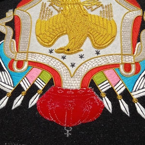 Op de afbeelding: Close-up van een geborduurd embleem met een gouden adelaarontwerp op een witte achtergrond. Het embleem heeft rode, blauwe en roze accenten, met witte veerd details. Zwarte en witte draden zijn zichtbaar.