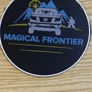 May include: A round black patch with a white border featuring the text "MAGICAL FRONTIER" in blue. The design includes a gray vehicle, blue mountains, a yellow compass, and a cartoon Bigfoot.