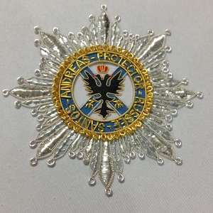 May include: A star-shaped embroidered patch featuring silver metallic thread rays. The center has a blue circular border with gold text reading ANDREAS-PROTECTOR-RUSSEE-SANTOS, surrounding a black double-headed eagle with a small red crown.