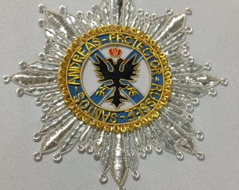 May include: A star-shaped embroidered patch featuring silver metallic thread rays. The center has a blue circular border with gold text reading ANDREAS-PROTECTOR-RUSSEE-SANTOS, surrounding a black double-headed eagle with a small red crown.