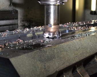 Może przedstawiać: Metalowe narzędzie frezarskie nacina duży, ciemnoszary blok stalowy, tworząc drobne metalowe wióry rozproszone po powierzchni. Cylindryczna głowica tnąca jest ustawiona pionowo i aktywnie obrabia metalowy przedmiot.