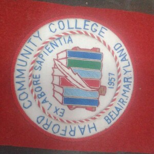 May include: A white embroidered patch with a red and white border. The patch features a blue and green book stack with a white feather and the text "COMMUNITY COLLEGE OF HARFORD COUNTY, LABORE SAPIENTIA EX, 1957".