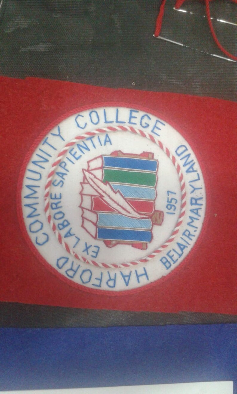 May include: A white embroidered patch with a red and white border. The patch features a blue and green book stack with a white feather and the text "COMMUNITY COLLEGE OF HARFORD COUNTY, LABORE SAPIENTIA EX, 1957".