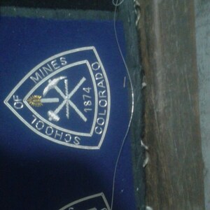May include: A white embroidered patch on a blue background. The patch is a triangle with the words "School of Mines" and "Colorado" on the sides and "1874" at the top. The patch also features a gold pickaxe and a gold torch.