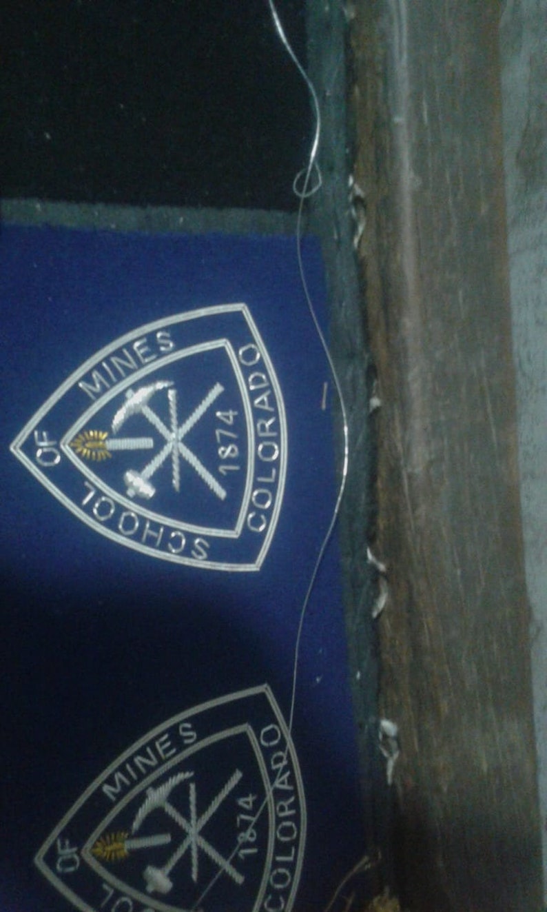 May include: A white embroidered patch on a blue background. The patch is a triangle with the words "School of Mines" and "Colorado" on the sides and "1874" at the top. The patch also features a gold pickaxe and a gold torch.