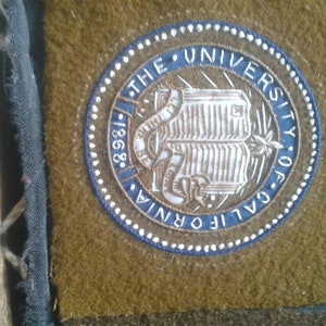 May include: A blue and white embroidered patch with the text "The University of California 1868" and a book with a snake wrapped around it.