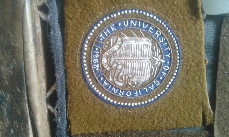 May include: A blue and white embroidered patch with the text "The University of California 1868" and a book with a snake wrapped around it.